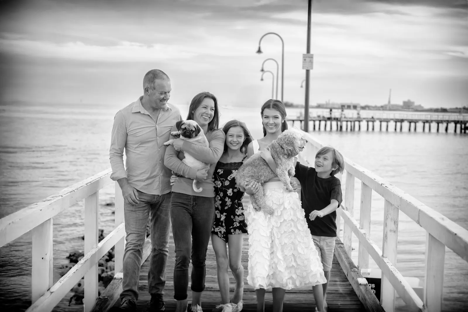 A family of five on a pier, holding a pug and a fluffy little dog, everyone laughing